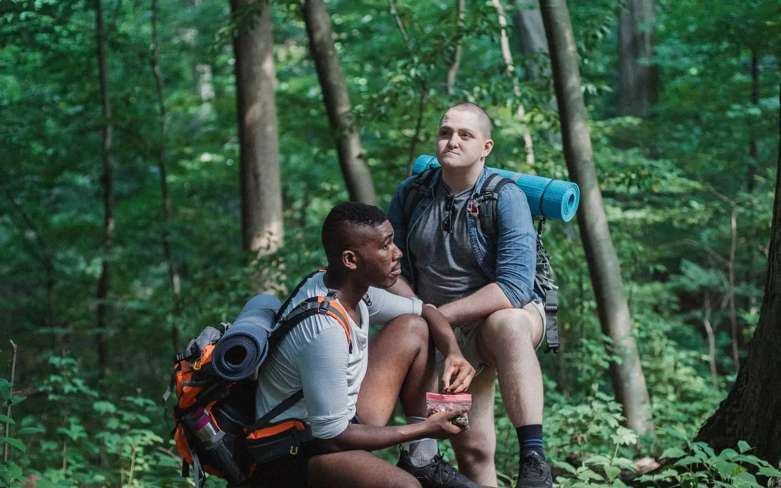 Two hikers with backpacks share a snack break in a green forest.