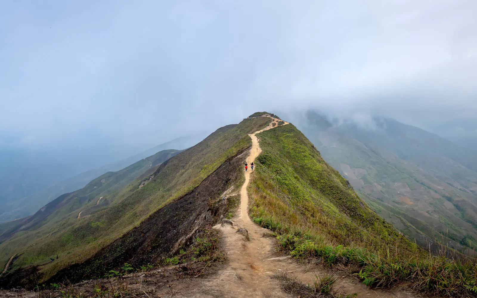 Two distant hikers cross an exposed mountain ridge as cloud covers the peaks ahead.