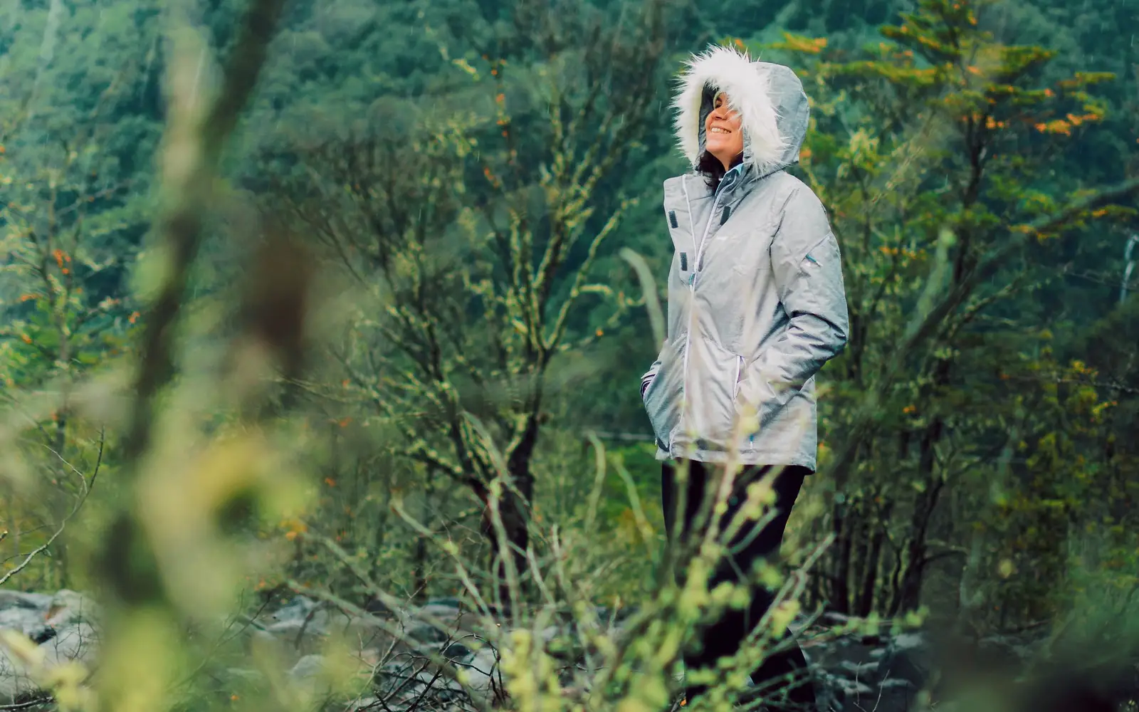 A hiker in a hooded weather layer stands on a wet green mountain trail in rain.