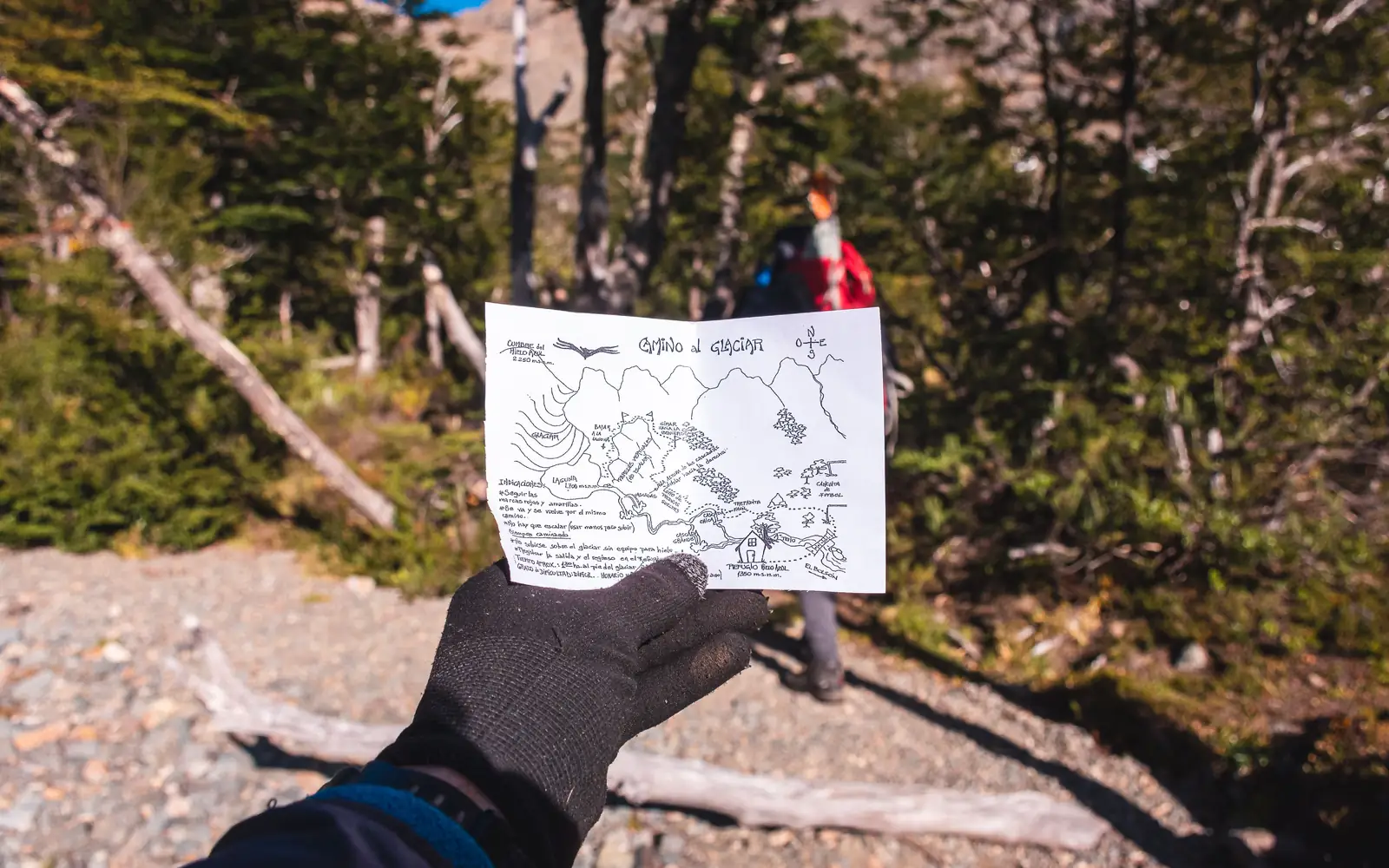 A gloved hiker holds a hand-drawn route map while another hiker waits on a forest trail behind.