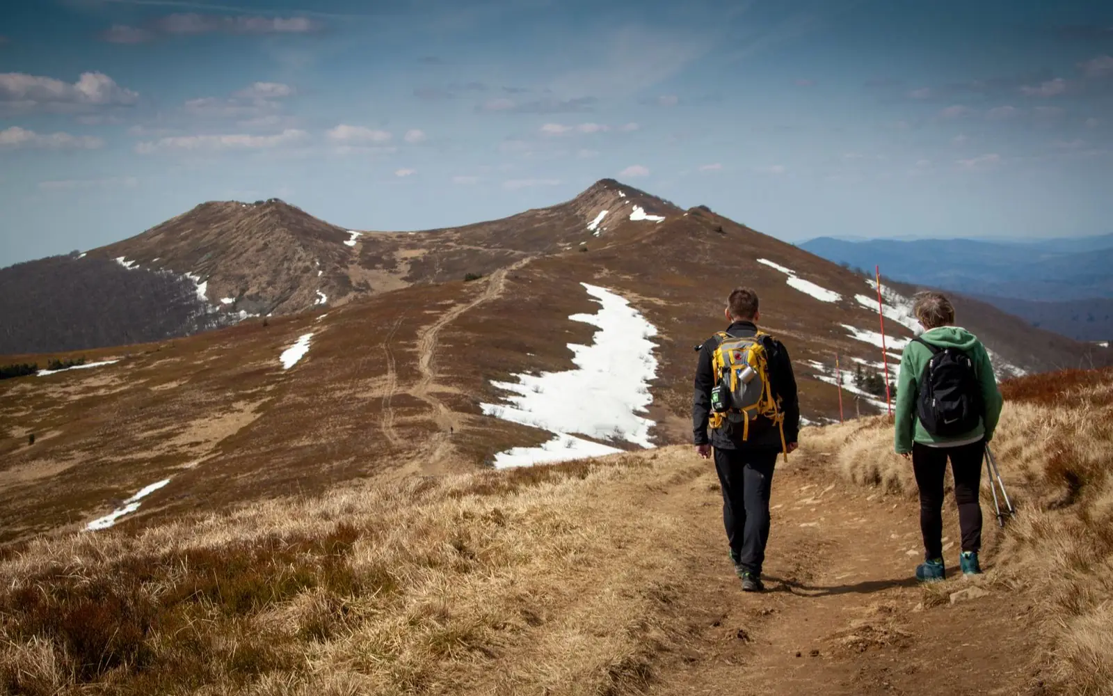 Two hikers travel steadily along a long mountain trail with spring snow patches.