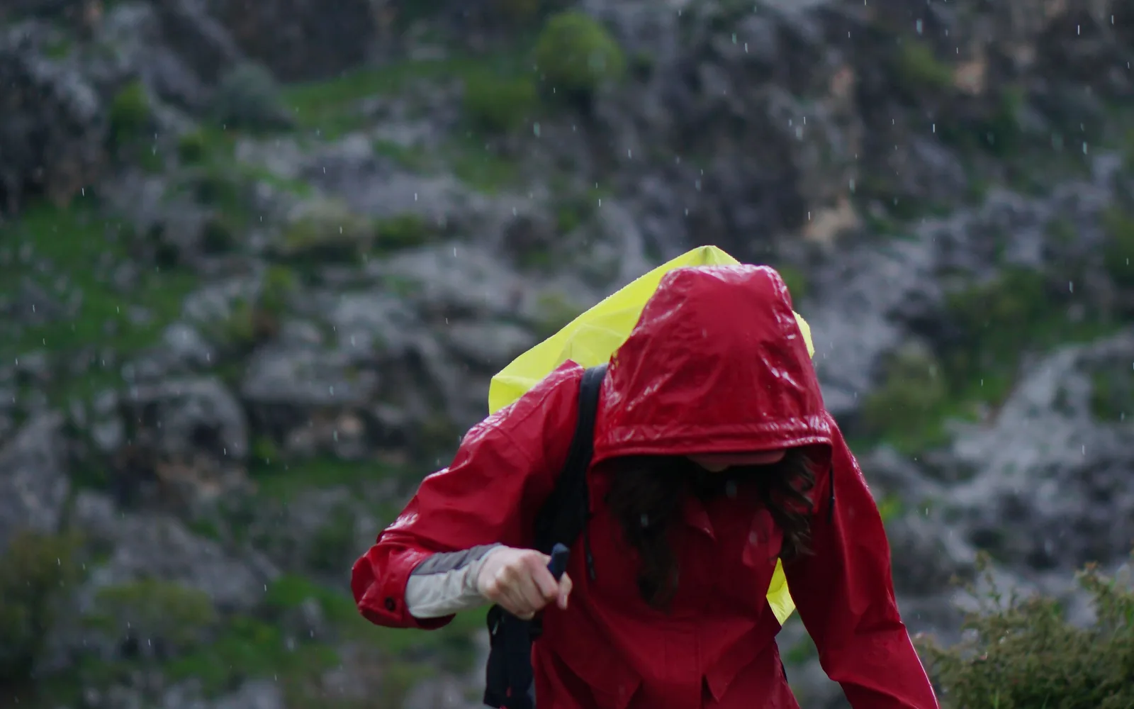 A hiker in a red rain jacket walks a rocky mountain trail under changing weather.