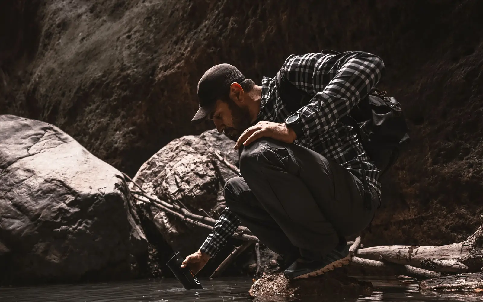A hiker crouches beside a rocky stream while filling a water-treatment container.