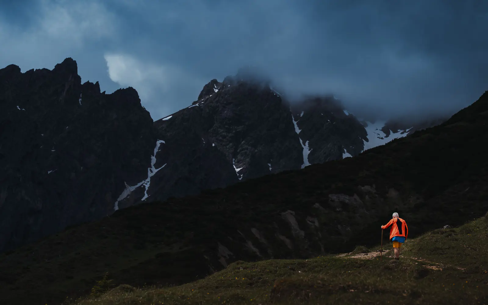 A lone hiker in a bright jacket approaches dark alpine peaks beneath low storm cloud.