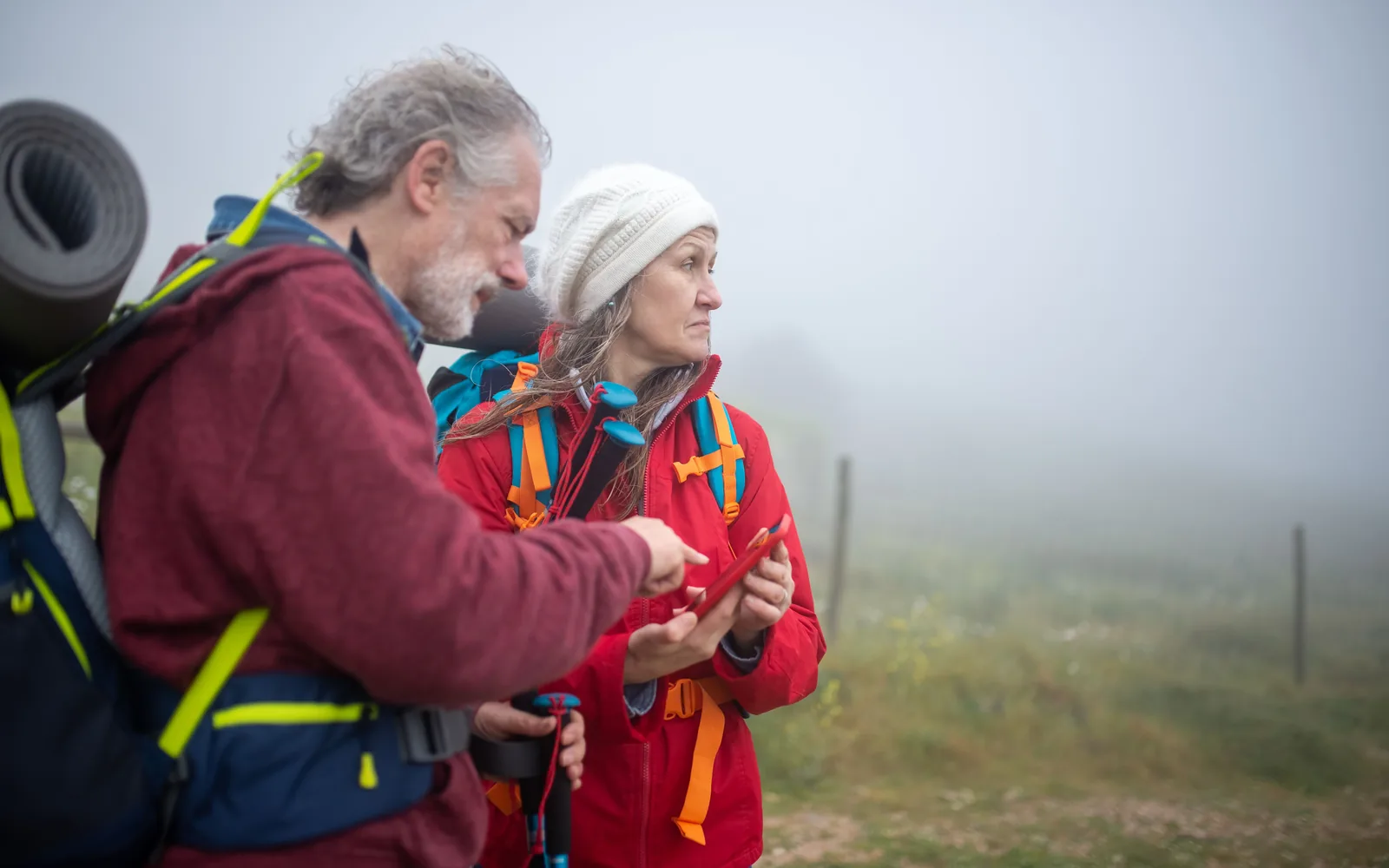Two hikers compare a smartphone map beside a foggy trail in open countryside.